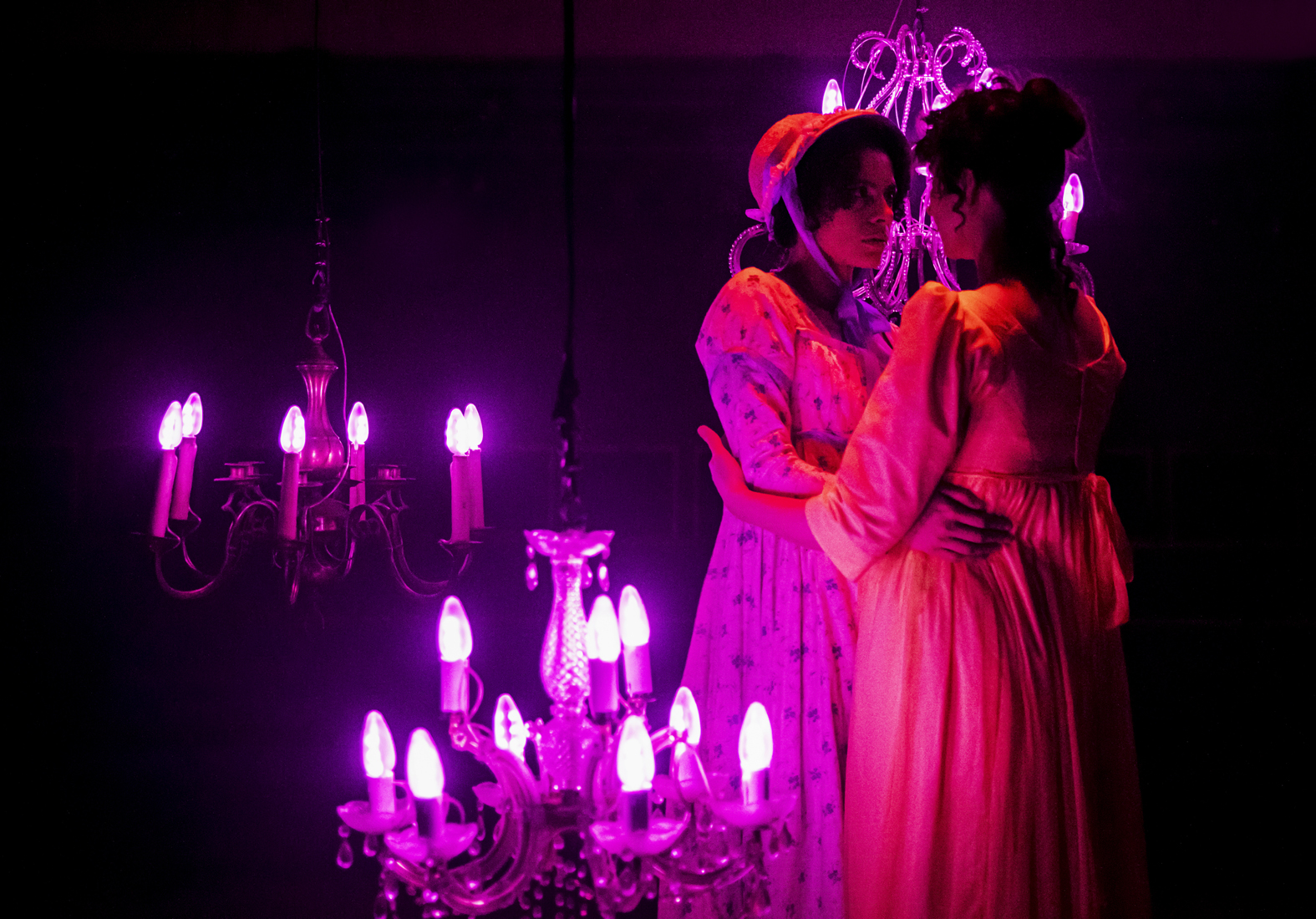 Two actors are hugging each other an dlooking into each others eyes. One is wearing a white dress with blue, floral print and a matching bonnet. The other is wearing a yellow dress. They are lit under a pink light and surrounding by lit chandeliers.