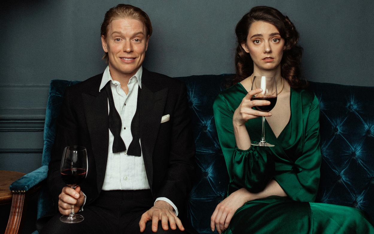 A fair man, and a brunette woman sit on a blue velvet sofa. He is dressed in a tux and white shirt with the bow tie undone and is smiling broadly; she is staring ahead clutching a wine glass of red wine, and wearing a green silk dress.