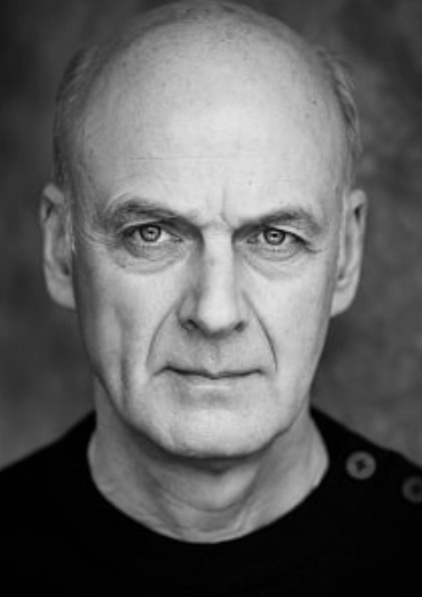 William Chubb's headshot: This image is in black and white. The actor wears a black jumper and has receding white hair.
