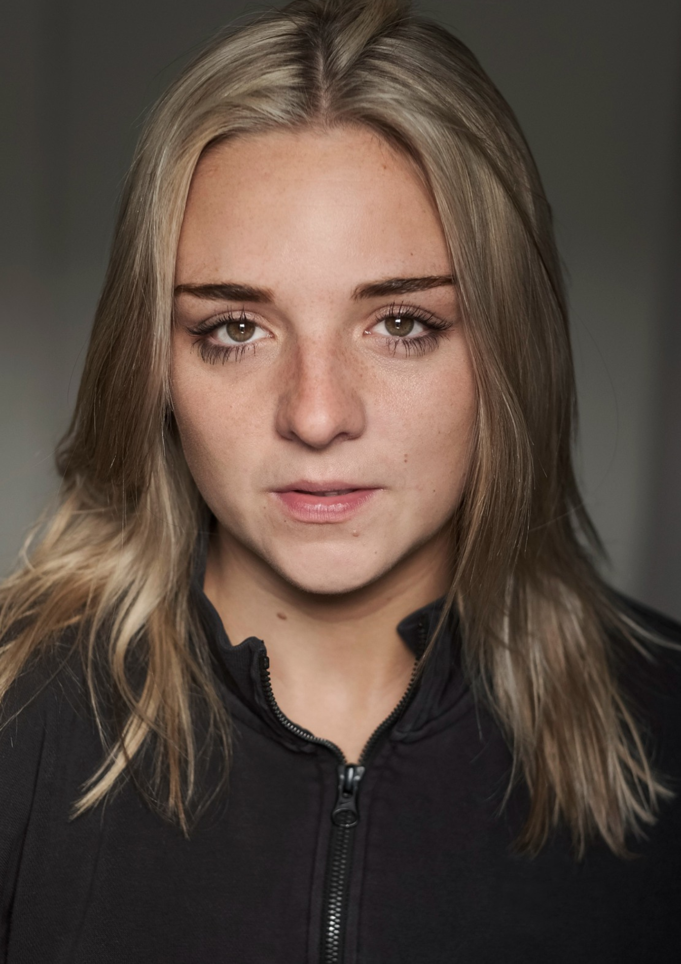 A headshot of a woman. She has long blonde hair and brown eyes. She wears a black zip-up jumper.