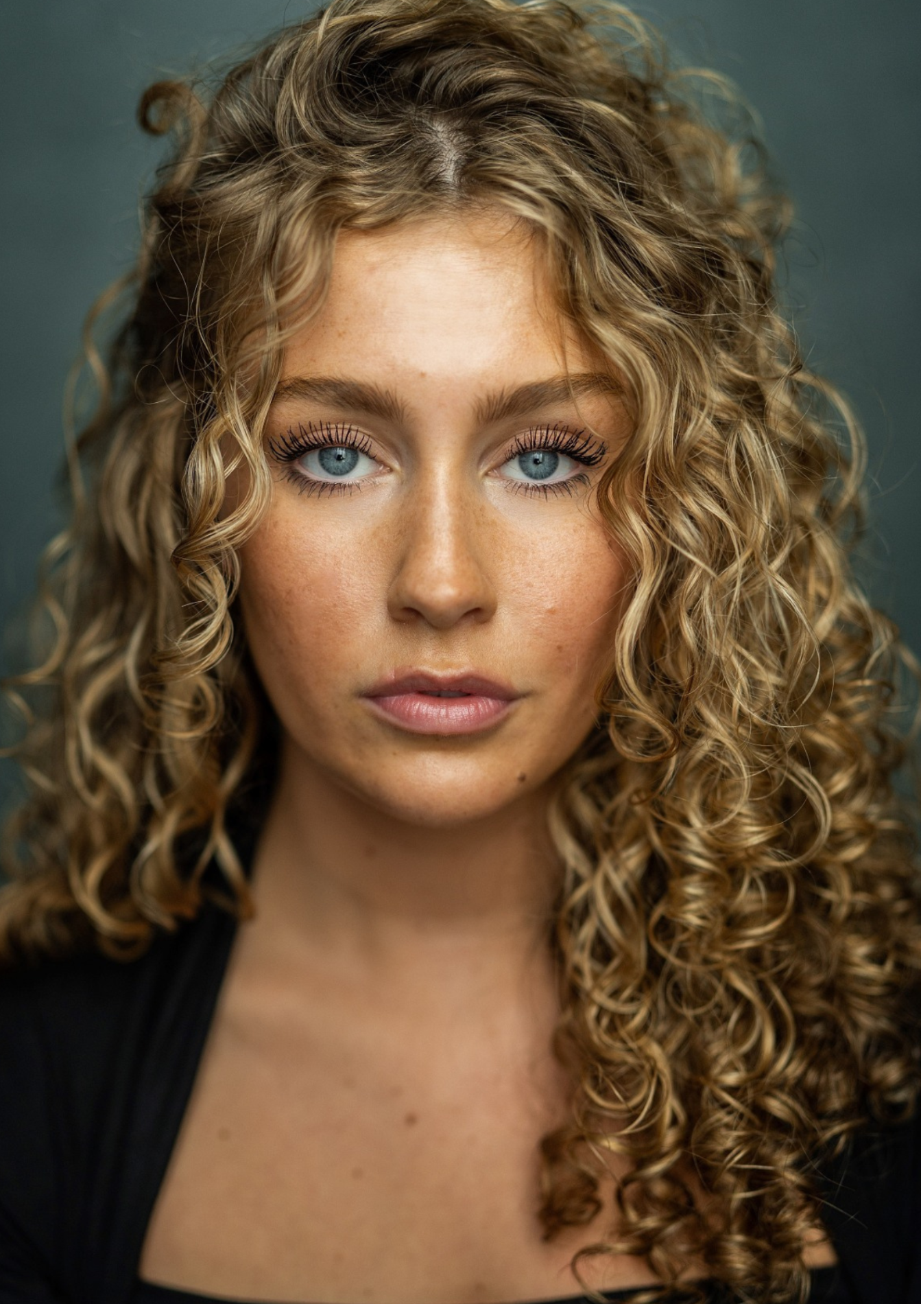 A headshot of a woman. She has blue eyes and long blonde curly hair. She wears a black blouse.