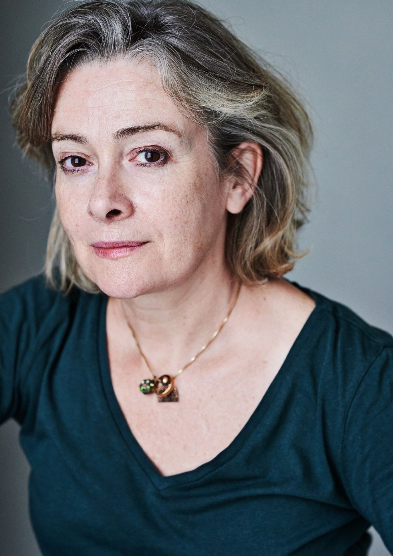 A woman with a grey and mousy bob. She has dark, brown eyes and is wearing a green, long-sleeved top and a gold necklace.