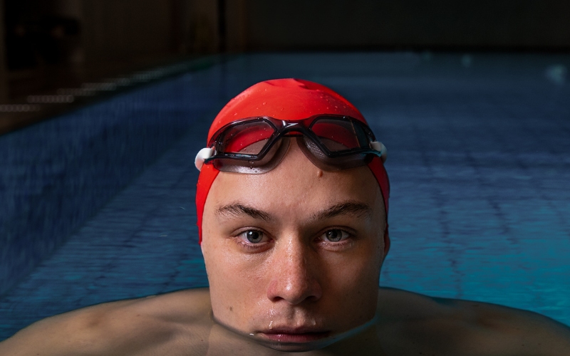 A man is seen in a swimming pool, submerged in water up to his chin. He wears a red swim cap with black swim goggles resting on his forehead. The length of the pool is visible behind him.