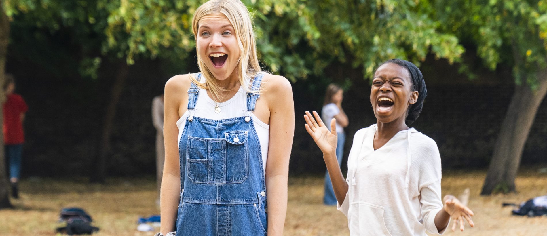 A student with shoulder length blond hair stands outside wearing denim overalls and a white t shirt. Her mouth is agape in surprise and delight. Behind her another student wearing a white jumper and blue joggers outstretches her arms and laughs.